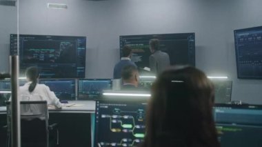 Dişi yazılım mühendisi bilgisayar başında elinde engellenmiş ağ ve gözetleme haritalarıyla oturuyor. Teknik destek uzmanları izleme odasında çalışıyor. Duvarda bir sürü büyük ekran var. Dolly vurdu..