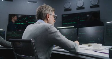 Olgun bir finans analisti gerçek zamanlı hisse senetleri ile PC 'de çalışır, döner ve kameraya bakar. Meslektaşlar, arka plandaki büyük ekranlardaki döviz piyasalarının grafiklerini analiz ederler. Yatırım konsepti. Portre.