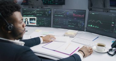 Afro-Amerikalı tüccar bilgisayar başında haber ve gerçek zamanlı hisse senetleriyle çalışıyor. Meslektaşlar arka planda döviz piyasalarının grafiklerini analiz ederler. Kripto para ticareti ve yatırım konsepti. Arka plan.