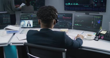 Afro-Amerikalı tüccar gerçek zamanlı sergilenen hisselerle bilgisayar başında çalışıyor. İş arkadaşları, arka plandaki büyük ekranlardaki döviz borsalarını analiz ediyor. Ticaret ve yatırım konsepti. Arkadan bak. Dolly vurdu..