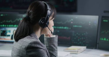 Bankada çalışan kulaklıklı kadın finansal analistin yakın çekimi. Gerçek zamanlı hisse senetleri, borsa grafikleri, grafikleri ve televizyon haberleri olan çoklu monitörlü bilgisayar. Analizler, kripto para ticareti