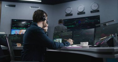 Elinde kalem olan tüccar bilgisayarında gerçek zamanlı stoklarla çalışıyor. Meslektaşlar, arka plandaki büyük ekranlardaki döviz piyasalarının grafiklerini analiz ederler. Kripto para ticareti ve yatırım kavramı.