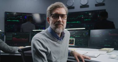 Olgun bir finans analisti gerçek zamanlı hisse senetleri ile PC 'de çalışır, döner ve kameraya bakar. Meslektaşlar, arka plandaki büyük ekranlardaki döviz piyasalarının grafiklerini analiz ederler. Yatırım konsepti. Portre.