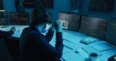 Elinde kalem olan tüccar bilgisayarında gerçek zamanlı stoklarla çalışıyor. Meslektaşlar, arka plandaki büyük ekranlardaki döviz piyasalarının grafiklerini analiz ederler. Ofis mavi ışıkla aydınlatılmış. Yatırım kavramı.