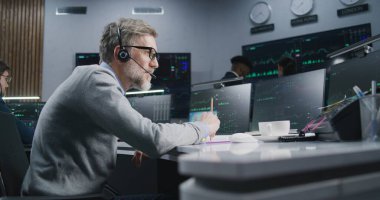 Kulaklık takan olgun bir erkek tüccar ofiste gerçek zamanlı hisse senedi olan çoklu monitörlü bilgisayarda çalışıyor. Meslektaşlar, büyük dijital ekranlarda döviz piyasalarının grafiklerini analiz ederler. Kripto para birimi ticareti ve analizi.