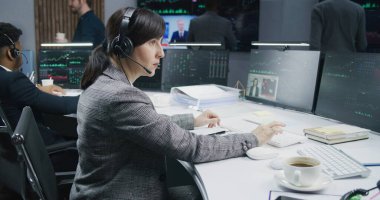 Dişi tüccar not defterine yazar ve bilgisayarda gerçek zamanlı sergilenen hisselerle çalışır. Meslektaşlar, arka plandaki büyük ekranlardaki döviz piyasalarının grafiklerini analiz ederler. Kripto para birimi ticareti ve yatırımı.