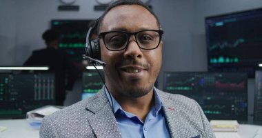 Kulaklık takan Afrikalı Amerikalı tüccar iş yerinde oturur, gülümser ve kameraya bakar. İş arkadaşları, arka plandaki büyük ekranlardaki döviz borsalarını analiz ediyor. Ticaret ve yatırım. Dolly vurdu. Portre.