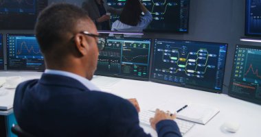 Yazılım programcısı izleme ofisinde engelleme ağı ve veri sunucusuyla bilgisayarın önünde oturur. IT teknik uzmanları büyük dijital ekranlarda canlı analiz çizelgeleri üzerinde çalışıyorlar..