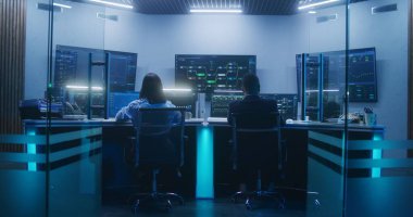 Geceleyin modern izleme ofisindeki veritabanı sunuculu bilgisayarlar üzerinde çalışan çeşitli bilişim uzmanlarının arka görüntüsü. Gerçek zamanlı analiz çizelgeleri ve engelleme ağına sahip büyük dijital ekranlar.