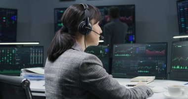 PC 'de çalışan kadın bir finans analisti gerçek zamanlı hisse senetleri gösteriyor, dönüyor ve kameraya bakıyor. Meslektaşlar arka planda döviz piyasalarının grafiklerini analiz ederler. Yatırım ve ticaret. Portre.