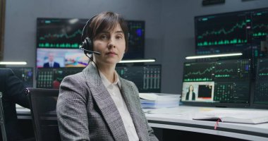 Kulaklık takan kadın tüccar bilgisayarda gerçek zamanlı hisse senetleriyle oturur ve kameraya bakar. Meslektaşlar, arka plandaki büyük ekranlardaki döviz piyasalarının grafiklerini analiz ederler. Yatırım ve ticaret. Portre.