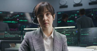 PC 'de çalışan kadın bir finans analisti gerçek zamanlı hisse senetleri gösteriyor, dönüyor ve kameraya bakıyor. Meslektaşlar arka planda döviz piyasalarının grafiklerini analiz ederler. Yatırım ve ticaret. Portre.