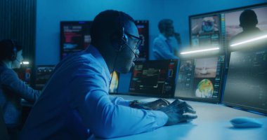 Kulaklıktaki Afro-Amerikan uçuş kontrol çalışanı komuta merkezindeki çoklu monitörlü bilgisayarda uzay görevini izliyor. Ekip büyük ekranlarda gösterilen başarılı uzay roketinin fırlatılmasından sonra el çırpıyor.