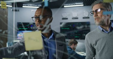 Çeşitli finansal analistlerden oluşan ekip cam duvara resim çiziyor, yatırım bankasındaki şirket iş stratejisini geliştiriyor ve analiz ediyor. Bilgisayarlar ve arka planda gerçek zamanlı hisse senetleri olan büyük dijital ekranlar.