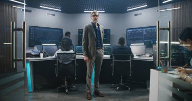 Olgun veri bilimci gözlük takar, izleme odasında durur ve kameraya bakar. Yazılım mühendisleri arka plandaki bilgisayarlarda çalışırlar. Canlı analiz beslemesi ile birden fazla büyük ekran.