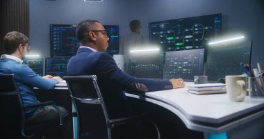 Çok ırklı bilişim uzmanları, ofislerinde veri sunucusu bulunan bilgisayarların önünde oturuyorlar. Yazılım mühendisi büyük dijital ekranlarda canlı analiz çizelgeleri ve engelleme ağıyla çalışıyor..