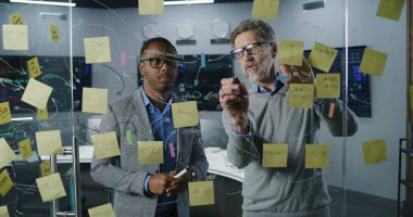 Çok ırklı finansal analistler yatırım bankasında cam duvara resim çiziyor, şirketin kripto iş stratejisini geliştiriyor ve analiz ediyor. Bilgisayarlar ve arka planda gerçek zamanlı hisse senetleri olan büyük dijital ekranlar