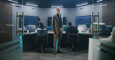 Olgun veri bilimci gözlük takar, izleme odasında durur ve kameraya bakar. Yazılım mühendisleri arka plandaki bilgisayarlarda çalışırlar. Canlı analiz beslemesi ile birden fazla büyük ekran.