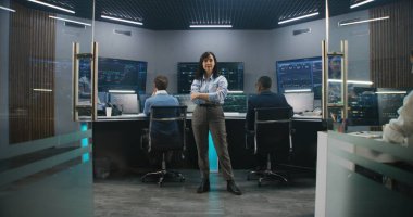 Dişi büyük veri bilimci izleme odasında duruyor ve kameraya bakıyor. Yazılım mühendisleri arka plandaki bilgisayarlarda çalışırlar. Gerçek zamanlı analiz çizelgeleri olan birden fazla büyük ekran. Dolly vurdu..