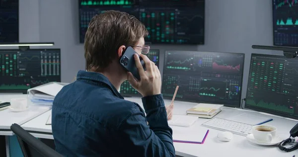 Male broker talks by phone, works in bank office on multi-monitor PC with real-time stocks, exchange market charts. Big digital screens with data. Investment, cryptocurrency trading and analytics.