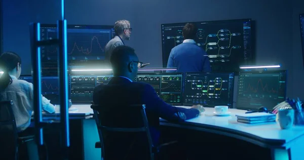 Multiethnic team of IT technical specialists and software programmers work with data server, analysis charts and blockchain network on computers and big digital screens in monitoring office at night.