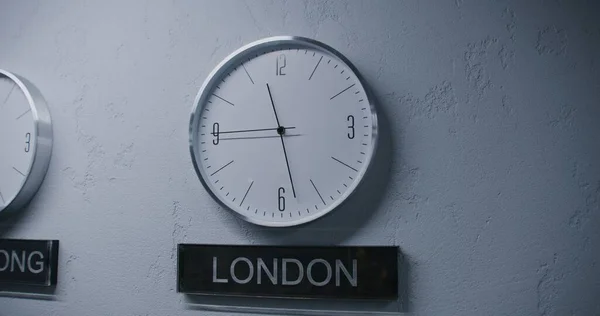 ランニングタイムポインタを備えたホワイトウォッチは 異なる都市のタイムゾーンを表示します モダンなデザインのオフィスでウォーキングウォールクロック 時計の下に書かれた大都市の名前 厳格なインテリア ドリーショット — ストック写真