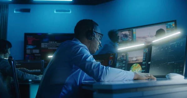 African American flight control employee in headset monitors space mission on multi-monitor computer in command center. Team clap hands after successful space rocket launch displayed on big screens.