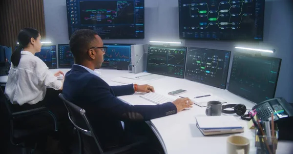 IT technical specialist sits in front of computer with blockchain network database in modern monitoring office. Team of software programmers work with real-time analysis charts on big digital screens.