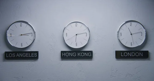 モダンなデザインのオフィスでランプによって照らされたランニングタイムポインタと白い壁の時計の静的なショット 壁時計の下に書かれた大都市の名前 異なる都市の時間を示す時計 — ストック写真