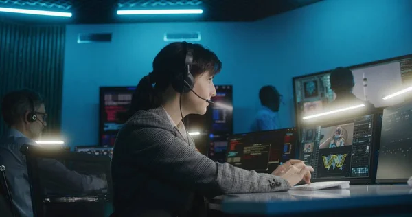Female flight control worker in headset sits in front of computer, monitors space mission in mission control center, looks at camera. Team watch space rocket lift off on big digital screens. Portrait.