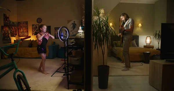 男は寝室でエレキギターを演奏する 若い女の子は リングランプと電話でソーシャルネットワークのためのダンスビデオを記録します 壁で区切られた2つの部屋やアパートのビュー 近所とライフスタイルのコンセプト — ストック写真