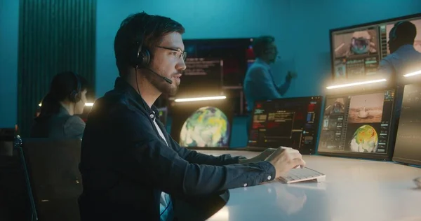 Flight control employee in headset sits in front of computers, monitors space mission in mission control center, looks at camera. Team watch space rocket launch on big digital screens. Portrait.