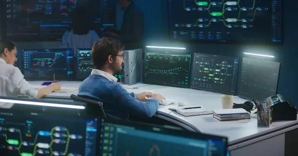 Software engineer works at computer in monitoring room. Technical support analyzes server data late night. Multiple big screens on the wall with displayed real-time analysis charts. Cyber security.