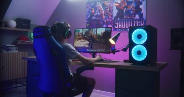 Genç oyuncu kişisel bilgisayarda üçüncü şahıs nişancıyı oynuyor. Kulaklıklı okul çocuğu evde online video oyunu seviyor. Masa, RGB LED lambasıyla aydınlatılmış. Evde oyun oynama kavramı. Arka plan.