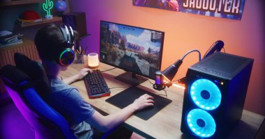 Genç oyuncu evdeki bilgisayarda oyun oynarken tankı kontrol ediyor. Bilgisayar monitörü çevrimiçi video oyunu canlı yayını ya da siber spor turnuvasıyla. Masa, RGB LED lambasıyla aydınlatılmış. Arka plan.