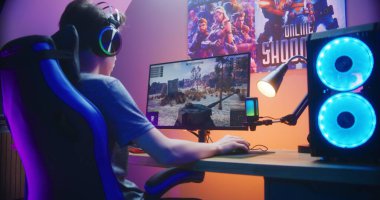 Genç oyuncu evdeki bilgisayarda oyun oynarken tankı kontrol ediyor. Bilgisayar monitörü çevrimiçi video oyunu canlı yayını ya da siber spor turnuvasıyla. Masa, RGB LED lambasıyla aydınlatılmış. Arka plan.