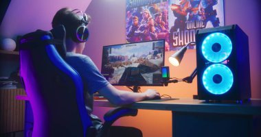 Genç oyuncu evdeki bilgisayarda oyun oynarken tankı kontrol ediyor. Bilgisayar monitörü çevrimiçi video oyunu canlı yayını ya da siber spor turnuvasıyla. Masa, RGB LED lambasıyla aydınlatılmış. Arka plan.