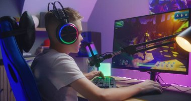 Kulaklıklı genç oyuncu takım arkadaşlarıyla konuşur ve evdeki bilgisayarda üçüncü şahıs nişancı ile oynar. Çevrimiçi video oyunu canlı yayın ya da esport yarışması. Ev bilgisayarında çok oyunculu PVP kavramı.