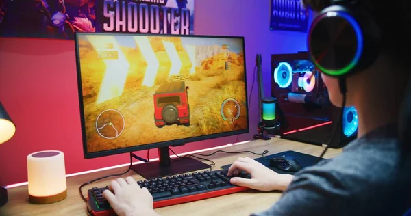 Young gamer plays in car driving simulator on PC at home. Computer monitor with displayed online video game live stream or cybersport championship. Desk illuminated by RGB LED strip light.