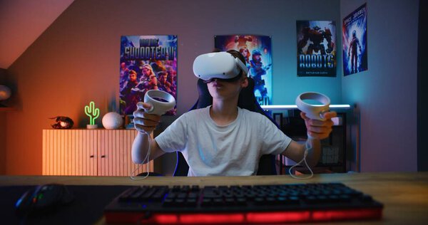 Young gamer in VR headset plays virtual online video game using wireless controllers. Boy enjoys eSports tournament. Room with warm neon LED lights. Gaming at home. POV from PC screen perspective.
