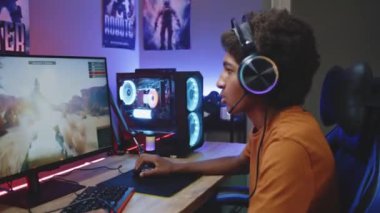 Üzgün Afro-Amerikalı kulaklıklı genç oyuncu çevrimiçi tank oyunu oynuyor PC 'de 3D oyun oynuyor, dönüyor ve kameraya bakıyor. Video oyunu yayını ya da eSports turnuvası. Evde oyun oynuyordum. Dolly vurdu. Yakınlaştır.