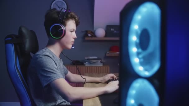 ヘッドフォンの若いゲーマーは自宅でコンピュータ上で再生されます 少年は振り向いてカメラに向かって話しかける Pcモニターに表示されるカーレースビデオゲーム 余暇を過ごす少年 ドリーは撃たれた サイドビュー — ストック動画
