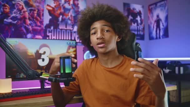 アフリカ系アメリカ人の10代のゲーマー ブロガーはマイクを使用してカメラで話す 親指を表示します ソーシャルネットワーク用のビデオゲームレビューを記録します ネオン居心地の良い部屋 背景にオンライン3DシューティングゲームとPc — ストック動画