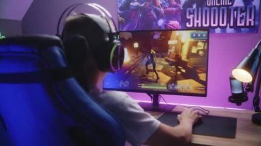 Genç çocuk kulaklık takar ve üçüncü şahıs nişancılık oynamaya başlar. Oyuncu ev bilgisayarında online video oyunu oynamayı seviyor. Masa, RGB LED lambasıyla aydınlatılmış. Ev konseptiyle oynamak. Arka plan.