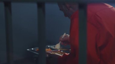 Hapishane hücresinde oturup yemek yiyen turuncu üniformalı yaşlı mahkumlara yakın çekim. Suçlu hapis cezasına çarptırıldı. Hapishane, gözaltı merkezi ya da ıslah evi. Metal parmaklıkların arasından ateş etmek.