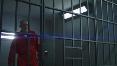 Turuncu üniformalı yaşlı suçlu parmaklıklı pencerenin yanında duruyor, hapishane hücresinin parmaklıklarına yaslanıyor ve kameraya bakıyor. Hapiste ya da gözaltı merkezinde. Kasvetli mahkum hapis cezasını çekiyor. Portre.