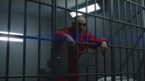 身穿橙色制服的老年囚犯把双手靠在金属棒上 罪犯在狱中服刑 在监狱 拘留中心或教养所的监牢后面站着面容憔悴的囚犯 — 图库视频影像