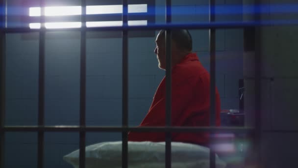 Пожилой Преступник Оранжевой Форме Сидит Тюремной Кровати Смотрит Окно Решетками — стоковое видео
