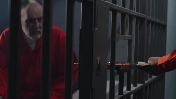 オレンジの制服を着た高齢の囚人が刑務所の独房に座っている 刑務所の警備員は金属棒を通して食物のトレイを与えます 有罪の犯罪者は懲役刑を言い渡される 刑務所又は拘置所の被収容者 — ストック動画