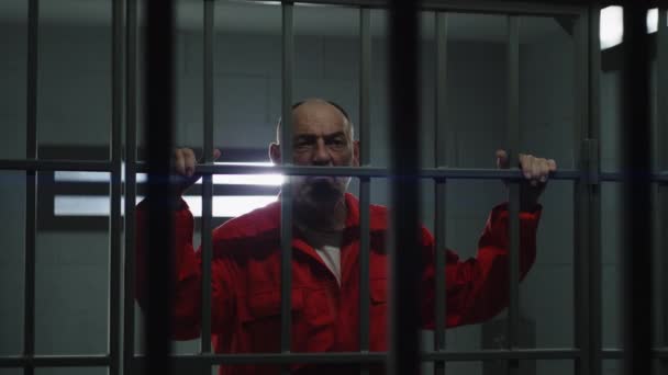 身穿橙色制服的老年囚犯手握金属棒 罪犯在狱中服刑 在监狱 拘留中心或教养所的监牢后面站着面容憔悴的囚犯 — 图库视频影像